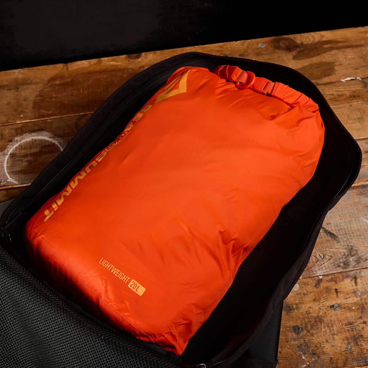 An orange Dry Sack from Sea to Summit, crafted with lightweight nylon, is neatly packed inside an open black backpack on top of a wooden surface. The sack is labeled with 