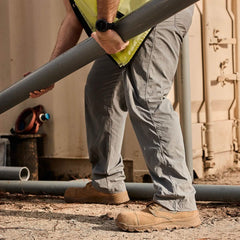 A person wearing gray trousers, a yellow safety vest, and the MACV-2 Safety Boot - Mid Top by GORUCK is lifting a large gray pipe near a container. Both hands are used to handle the pipe, suggesting a construction or maintenance context. A red valve is visible in the background.