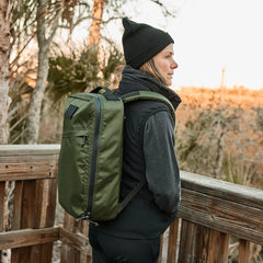 Person wearing black beanie and vest with olive green GORUCK backpack on wooden trail overlooking natural landscape at sunset