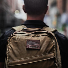A person faces away from the camera, displaying the GORUCK GR1 USA - Heritage backpack made from waxed duck canvas. The visible GORUCK logo patch adds to its appeal. A blurred city street behind them emphasizes the durable and stylish characteristics of this rucksack.