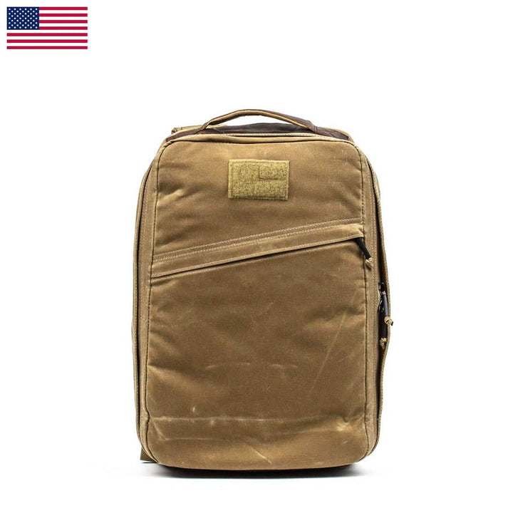 The GORUCK GR1 USA - Heritage rucksack, made from waxed duck canvas, stands upright against a white background with its tan fabric enhanced by a top handle and a front zippered pocket. Its durability is bolstered by the Martexin Wax finish, and an American flag is featured in the top left corner.