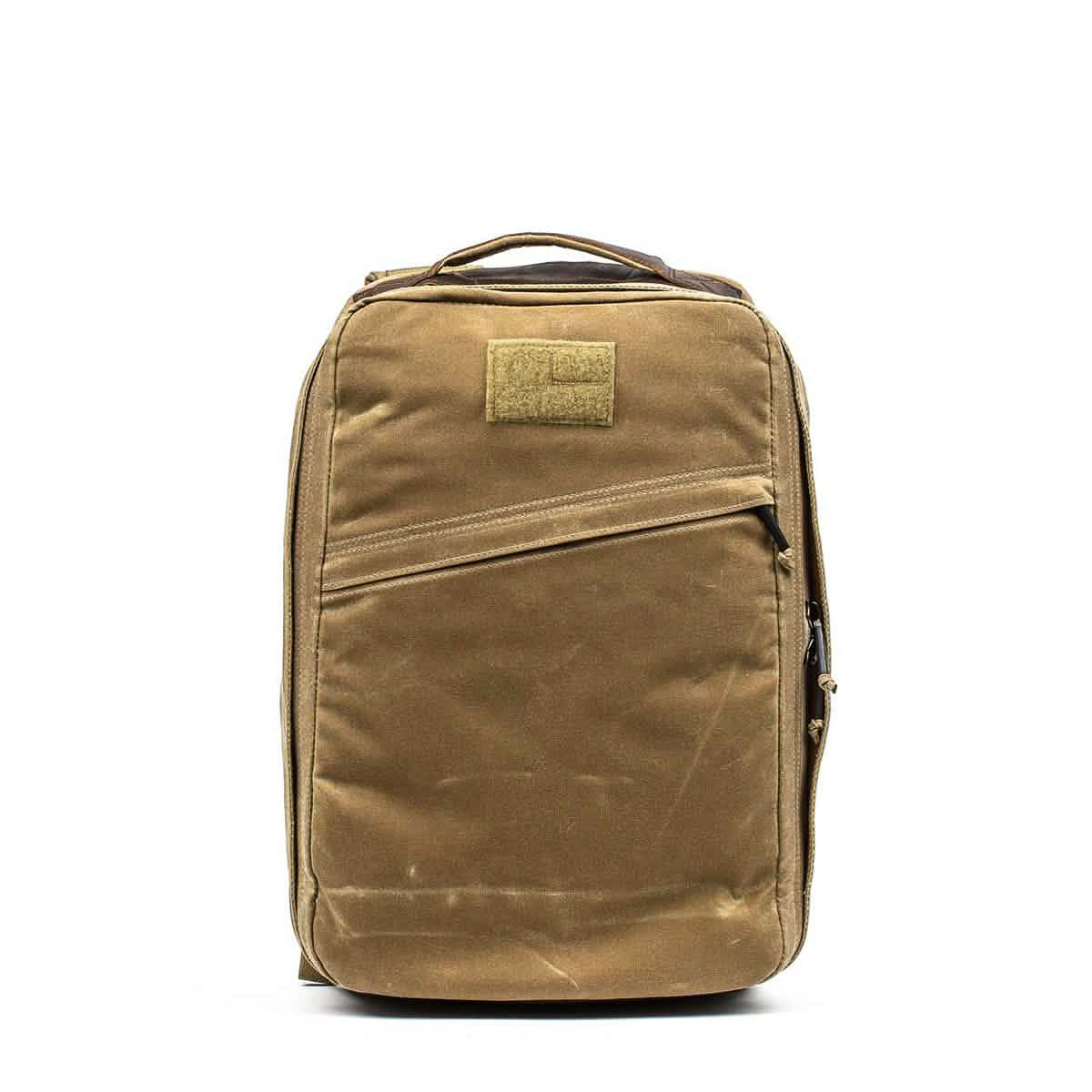 The GR1 USA - Heritage Waxed Canvas is a tan backpack featuring a top handle and a diagonal front zipper pocket, displayed against a white background.
