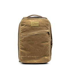 The GR1 USA - Heritage Waxed Canvas is a tan backpack featuring a top handle and a diagonal front zipper pocket, displayed against a white background.