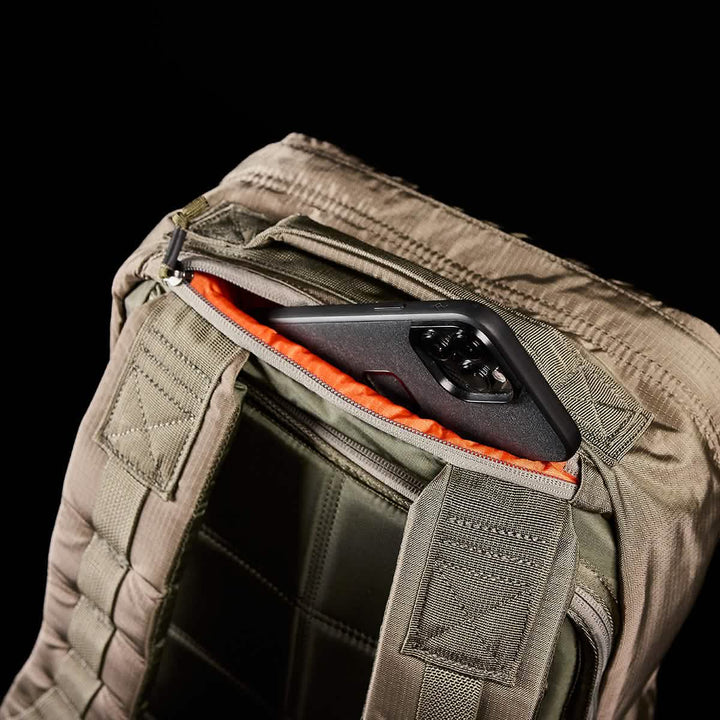 A smartphone is partially tucked into the top pocket of a khaki green GR1 USA - Ripstop ROBIC® rucksack by GORUCK, featuring quilted padding. The black phone case is visible against the bright orange, ultra-lightweight, water-resistant interior lining of the pocket.