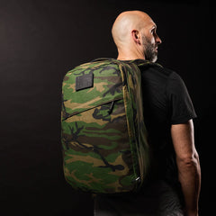 Bald man in a black shirt and large GR1 USA - Heritage Woodland Camo M81 backpack, shown sideways against a dark background.