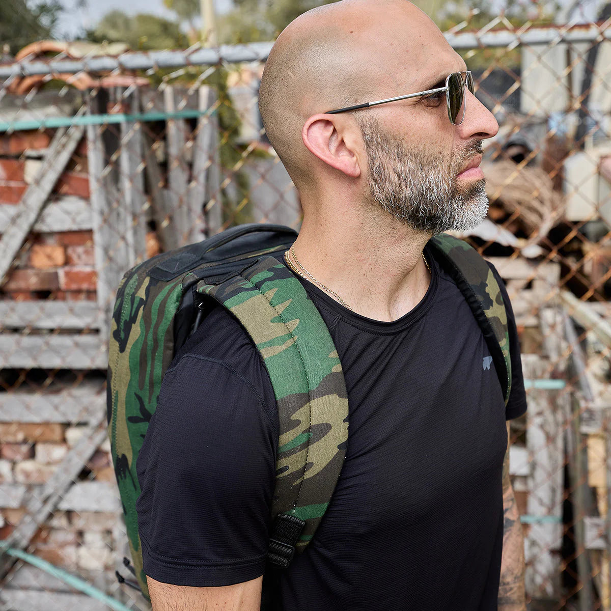 Bald man with sunglasses and a beard, dressed in a black shirt, stands outdoors carrying the GR1 USA - Heritage Woodland Camo M81 backpack.