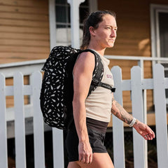 Woman wearing tan GORUCK tank top and black shorts with patterned black GORUCK backpack walking outdoors by white picket fence
