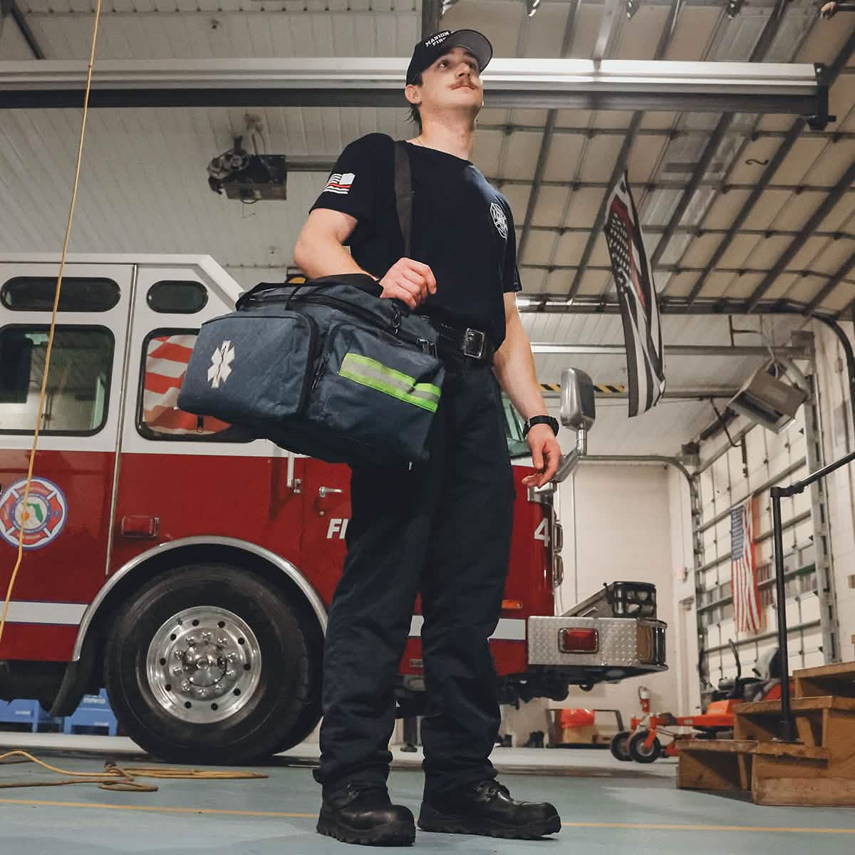 A firefighter, who meets Special Forces standards, stands in a fire station holding a large emergency medical bag. Wearing his black uniform and the GORUCK MACV-2 Safety Boot - High Top, he is flanked by a nearby fire truck and an American flag. The brightly lit station exemplifies organization and readiness.