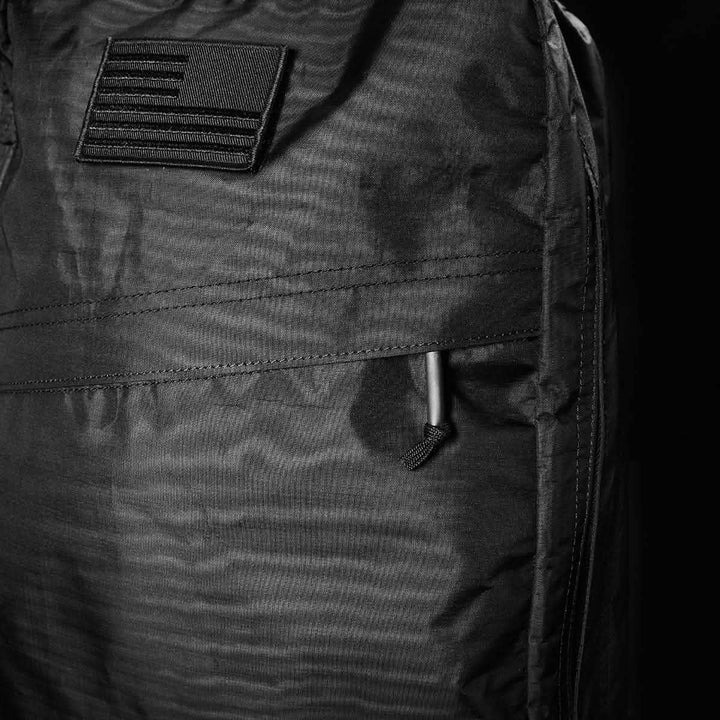 A detailed view showcases the GR1 USA - Dyneema jacket by GORUCK, featuring a black fabric design with a monochrome American flag patch on the shoulder. Constructed from durable Dyneema®, the texture is emphasized with visible stitching and an accentuated side zipper pull. The lighting gently casts soft shadows across the surface, enhancing its characteristics.