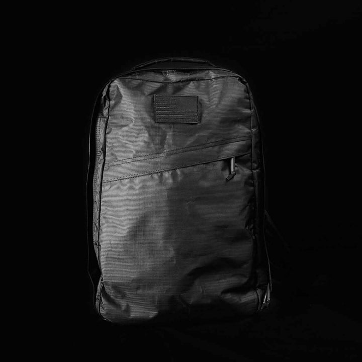A sleek black GORUCK GR1 USA - Dyneema rucksack with a front zipper pocket, set against a dark background. The material exhibits a slightly textured appearance, accompanied by a small rectangular patch near the top.