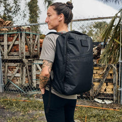 Person with tattoos wearing a black GORUCK backpack outdoors near a chain-link fence and stacked wood