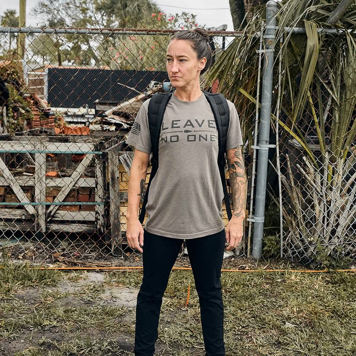 A person stands confidently in front of a chain-link fence and foliage, wearing a t-shirt with the words LEAVE NO ONE and black pants, complemented by a GORUCK GR1 USA - Dyneema rucksack. Their hair is tied back, revealing tattooed arms, while wooden crates and bricks are visible in the background.