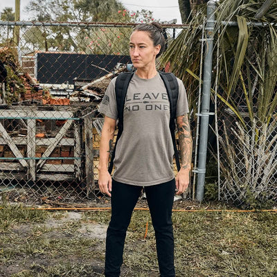 A person stands confidently in front of a chain-link fence and foliage, wearing a t-shirt with the words LEAVE NO ONE and black pants, complemented by a GORUCK GR1 USA - Dyneema rucksack. Their hair is tied back, revealing tattooed arms, while wooden crates and bricks are visible in the background.
