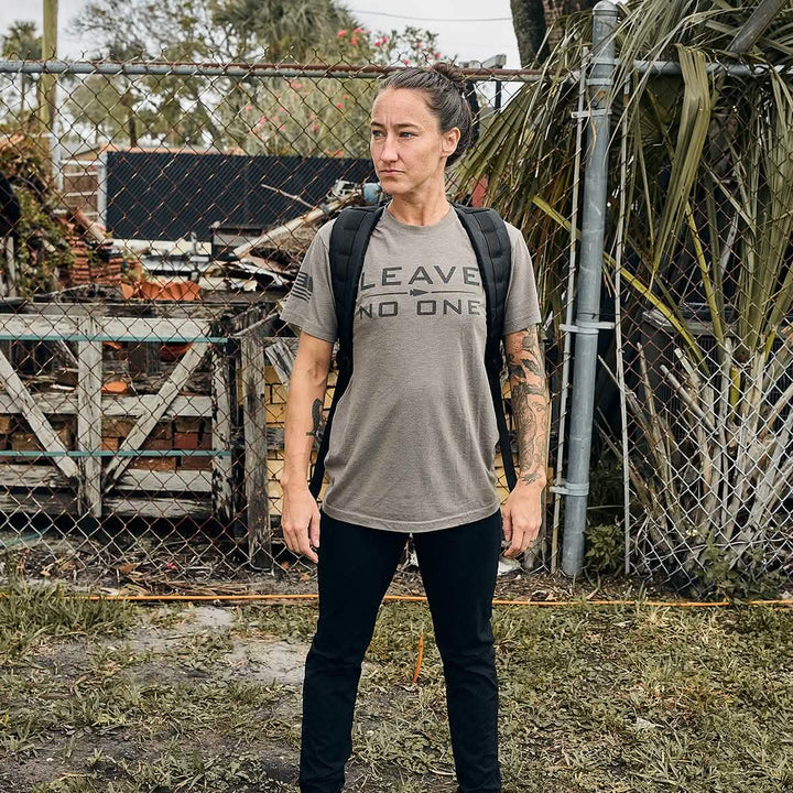 A person stands confidently in front of a chain-link fence and foliage, wearing a t-shirt with the words LEAVE NO ONE and black pants, complemented by a GORUCK GR1 USA - Dyneema rucksack. Their hair is tied back, revealing tattooed arms, while wooden crates and bricks are visible in the background.