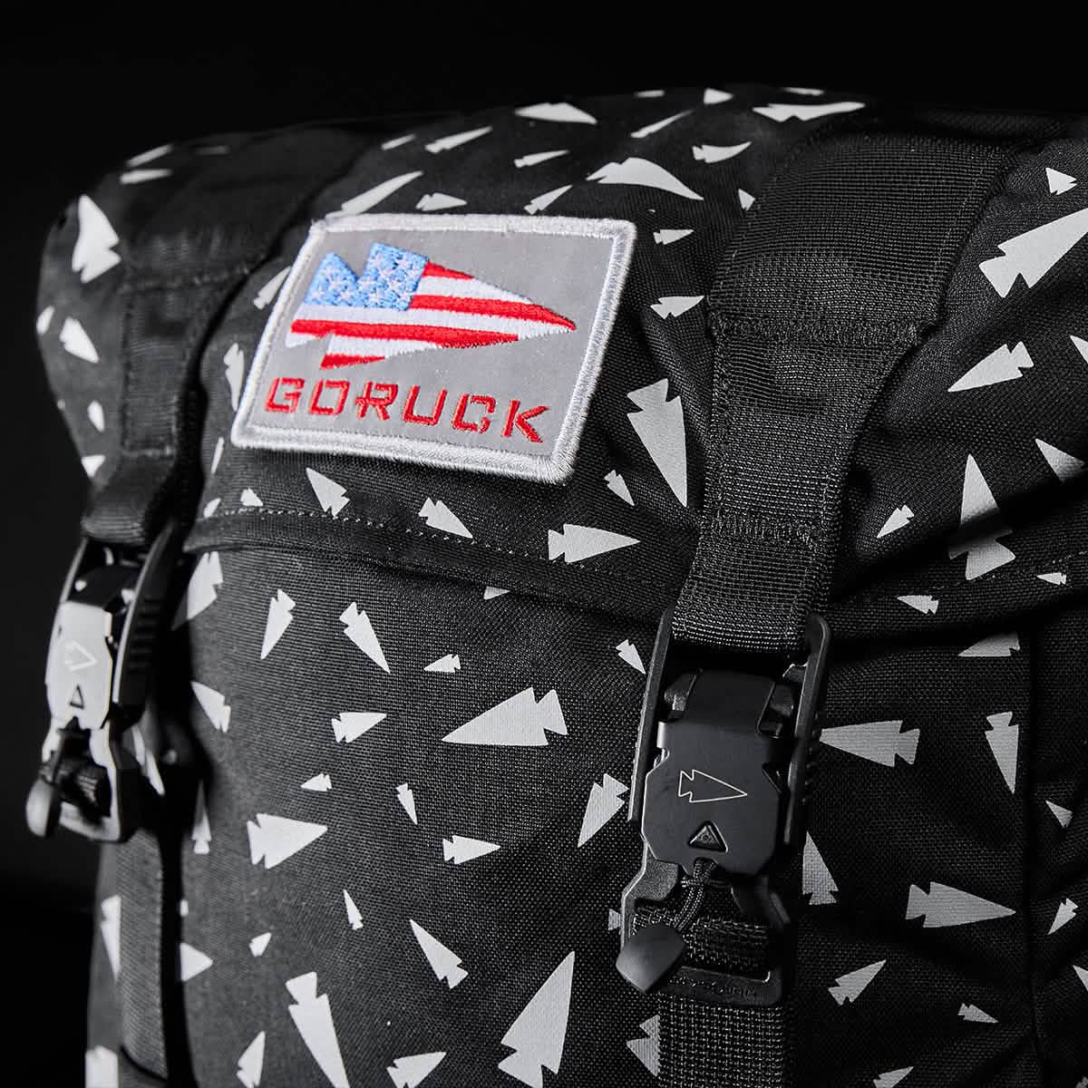 Close-up of the GORUCK M23 Spearhead Shrapnel rucksack featuring a black design with white geometric patterns. The front flap displays a bold patch of the American flag along with the 