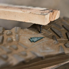 Close-up of a MACV-2 Safety Boot - Mid Top by GORUCK, showing a large nail piercing through a wooden board and lodged into the tread. The scene highlights the potential danger of stepping on sharp objects. Its composite toe and textured pattern, along with some dirt, hint at its rugged past.