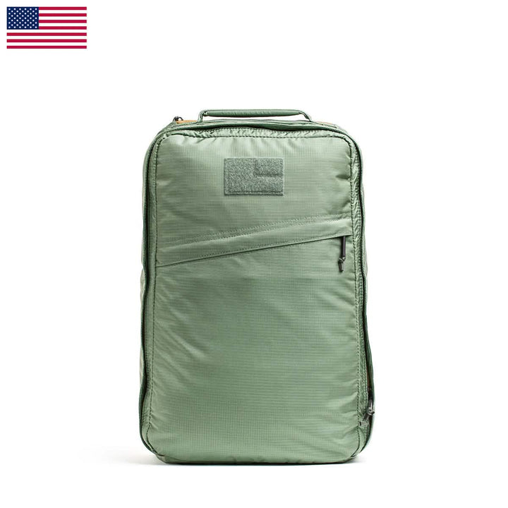The GR1 USA - Ripstop ROBIC® by GORUCK is a lightweight green rucksack featuring a front zipper pocket and a small patch area on top. Its water-resistant design contrasts with the white background, highlighted by a small USA flag in the top left corner.