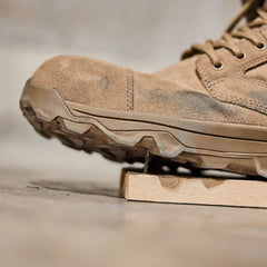 A close-up of a lightly worn MACV-2 Safety Boot - Mid Top from GORUCK placed on a wooden board. The boot features rugged soles and sturdy laces, suggesting Special Forces standards for durability and outdoor use. The background is out of focus, emphasizing the boot's texture and detail.