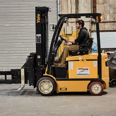 A man wearing GORUCK's MACV-2 Safety Boot - Mid Top skillfully operates a yellow Yale forklift inside a warehouse with a closed roller door in the background. Focused ahead, he expertly steers the vehicle on the concrete floor, precisely navigating various warehouse items around him with precision akin to Special Forces standards.