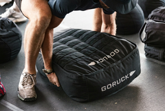 Person performing loaded carries with a large black Sand Tombstones sandbag in a gym, wearing athletic clothing and sneakers.