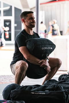 A man in a squat holds a large loadable Sand Tombstones bag labeled "GORUCK" outdoors, with gear bags around him.
