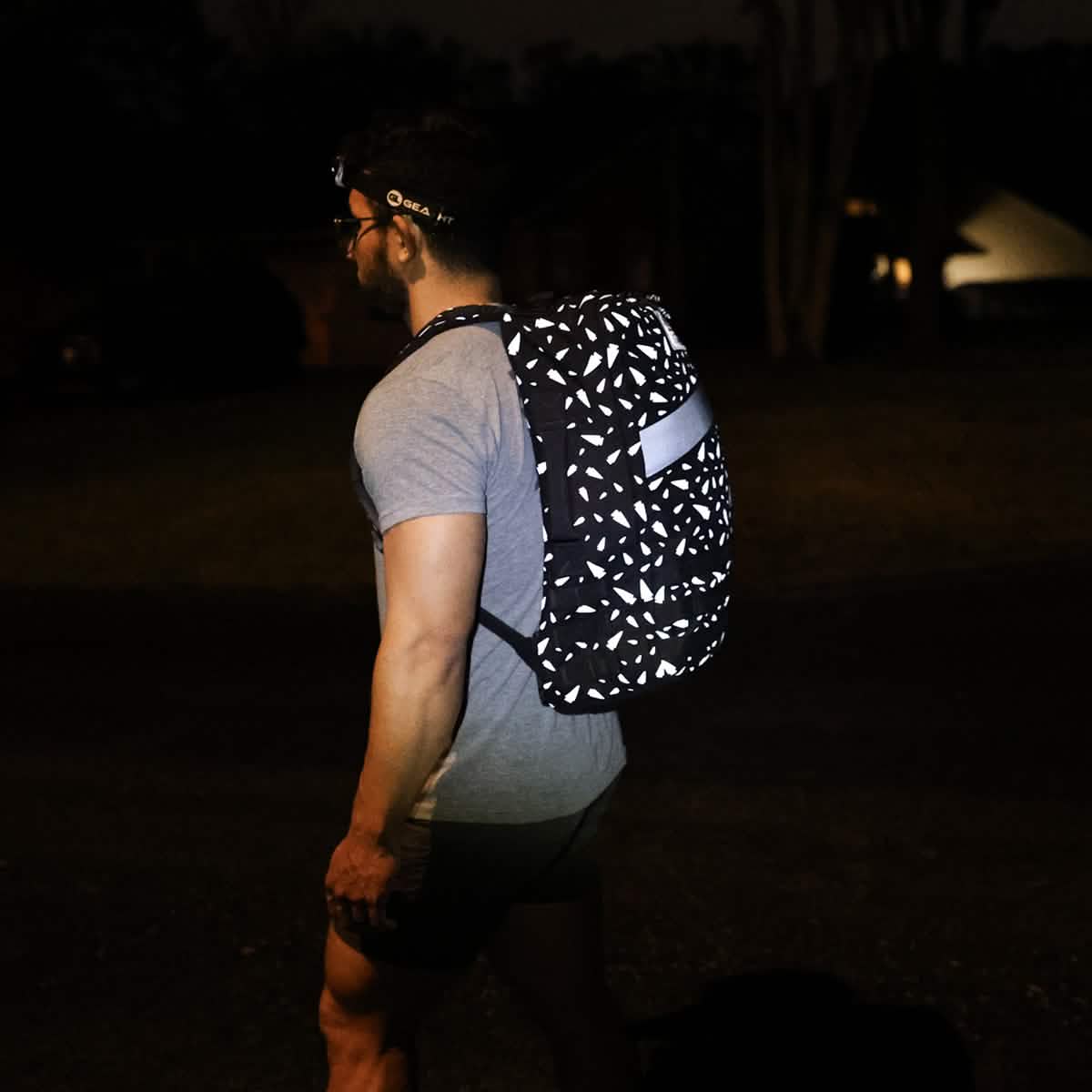 A man stands outside at night, facing away from the camera, wearing a headlamp and the Rucker 4.0 - Reflective Spearhead backpack.