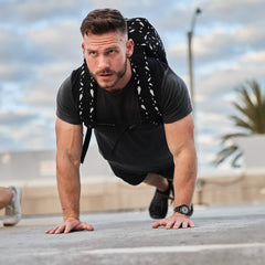 A bearded man does push-ups outdoors, wearing a black T-shirt and the Rucker 4.0 - Reflective Spearhead rucksack, showcasing his dedication to rucking.