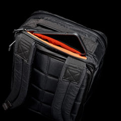 Black GORUCK rucksack with padded straps and an open top pocket revealing bright orange lining and a smartphone