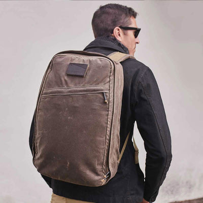 A person wearing sunglasses and a black jacket carries the GR1 USA - Heritage rucksack by GORUCK, crafted from waxed duck canvas with a Martexin Wax finish. The background is a plain light color.