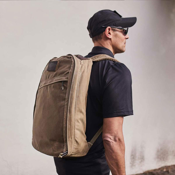 An individual sporting sunglasses and a black cap strolls along with a large GR1 USA - Heritage rucksack by GORUCK, crafted from waxed duck canvas. They are dressed in a black shirt against a light-colored wall.