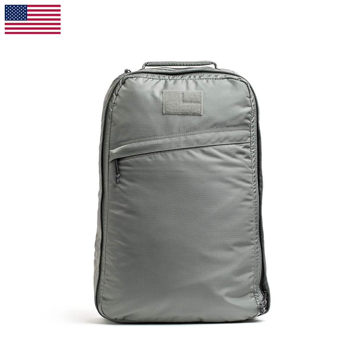 The GORUCK GR1 USA - Ripstop ROBIC® backpack is a gray ultra-lightweight, water-resistant bag with a minimalist design. It features a single front zipper pocket, a small patch on top, and the United States flag displayed in the top left corner. For added convenience, it also includes a handle.