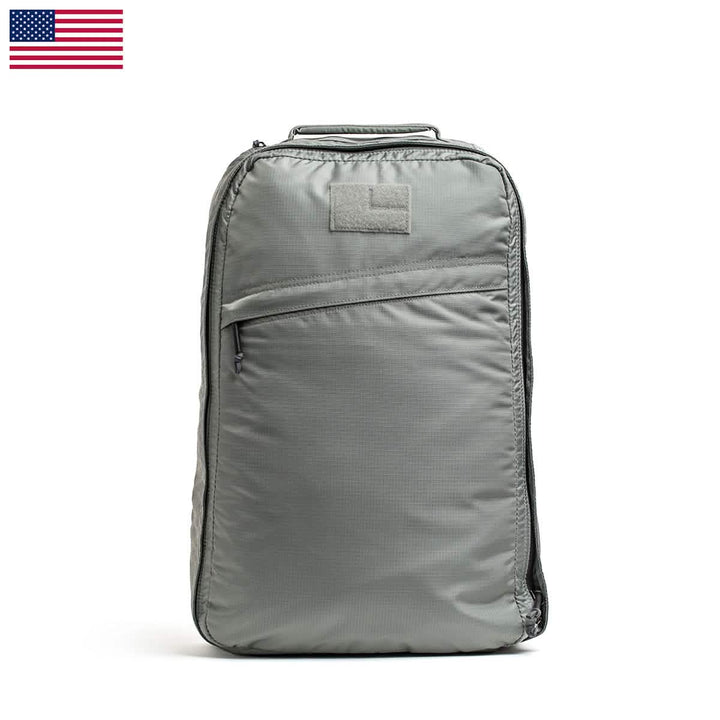 The GORUCK GR1 USA - Ripstop ROBIC® backpack is a gray ultra-lightweight, water-resistant bag with a minimalist design. It features a single front zipper pocket, a small patch on top, and the United States flag displayed in the top left corner. For added convenience, it also includes a handle.