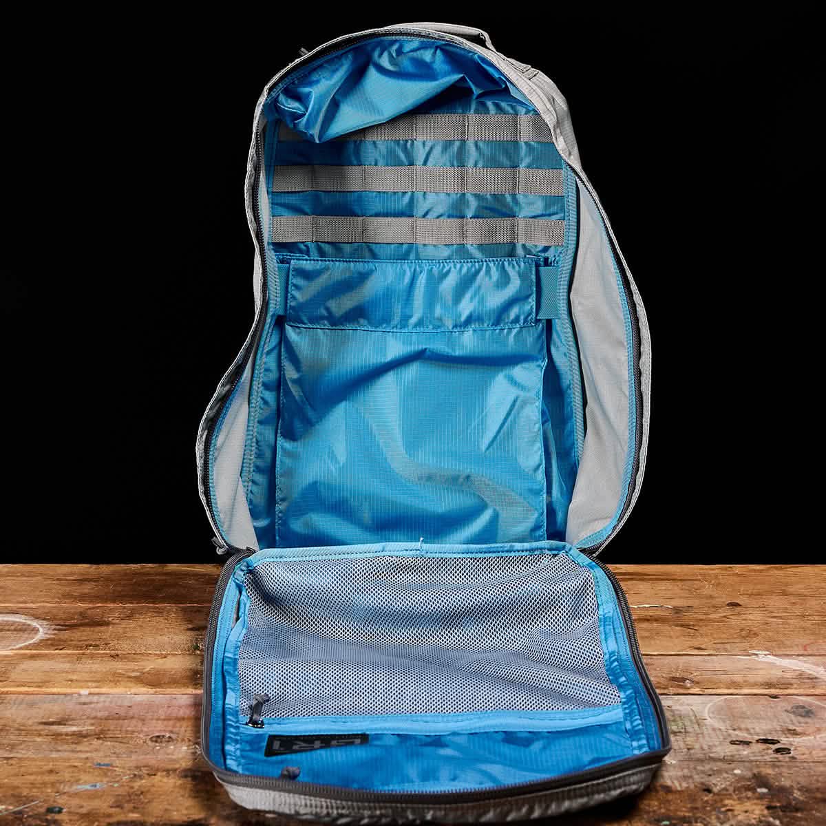 The GR1 USA - Ripstop ROBIC® by GORUCK, an ultra-lightweight rucksack in open gray with a blue interior, is displayed on a wooden surface against a black background. It features mesh pockets and organized compartments designed for efficient storage.