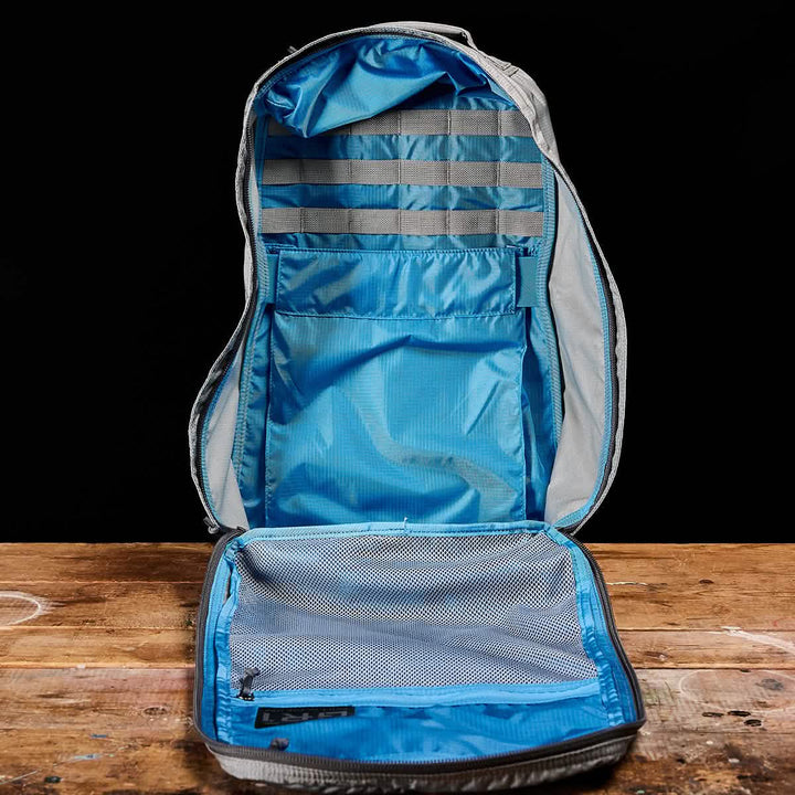 The GR1 USA - Ripstop ROBIC® by GORUCK, an ultra-lightweight rucksack in open gray with a blue interior, is displayed on a wooden surface against a black background. It features mesh pockets and organized compartments designed for efficient storage.