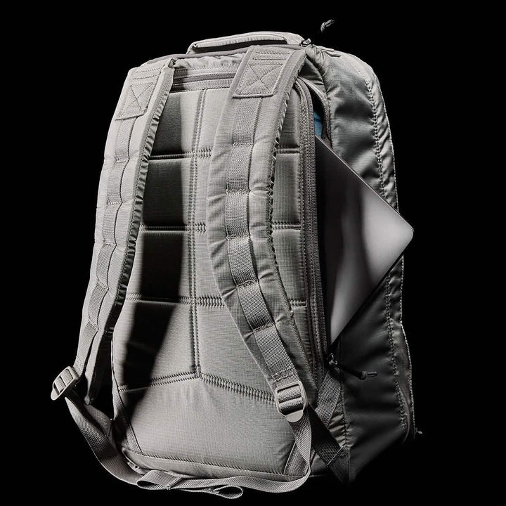 The GR1 USA - Ripstop ROBIC® by GORUCK is featured against a black background, showcasing its gray color and ultra-lightweight design. Padded shoulder straps enhance comfort as a laptop partially emerges from the back compartment, highlighting the backpack's metallic surface and water-resistant features.