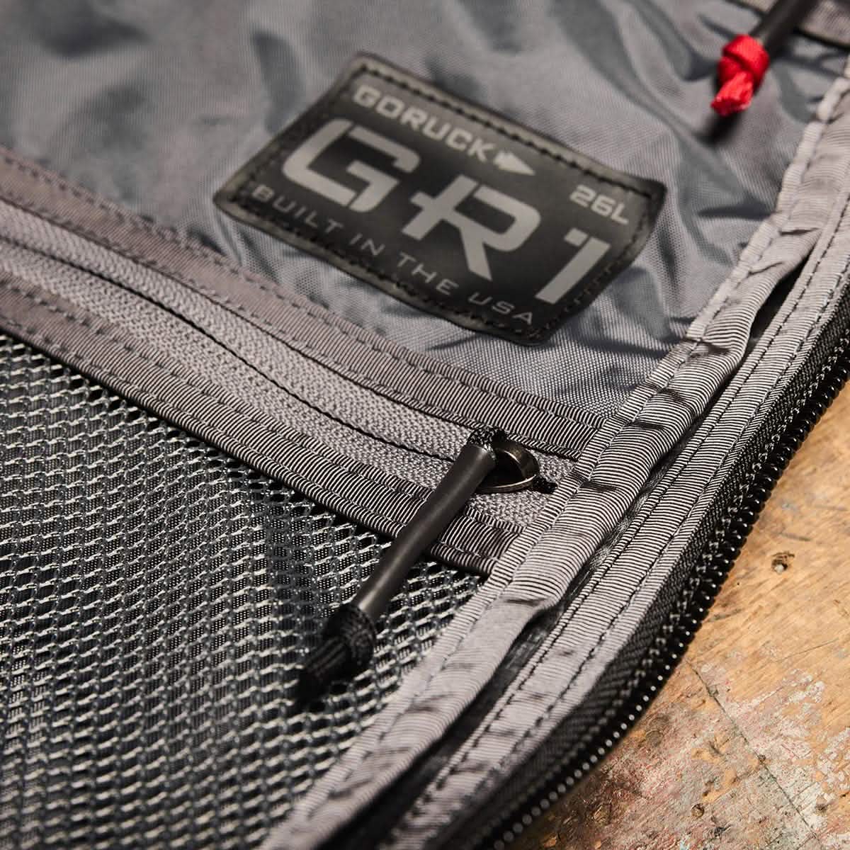 Close-up of a gray fabric rucksack showcasing a GORUCK GR1 USA - Dyneema patch and a mesh pocket. Visible zippers with black pulls add detail. Set against a rustic wooden surface, the background perfectly highlights the stylish utility of the GR1 design by GORUCK.