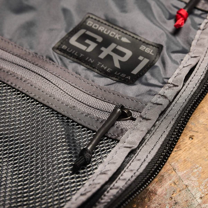 Close-up of a gray fabric rucksack showcasing a GORUCK GR1 USA - Dyneema patch and a mesh pocket. Visible zippers with black pulls add detail. Set against a rustic wooden surface, the background perfectly highlights the stylish utility of the GR1 design by GORUCK.