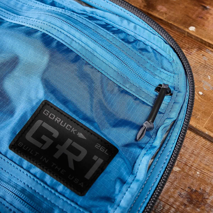 A close-up of a blue GORUCK GR1 USA - Ripstop ROBIC® rucksack interior reveals a mesh pocket with a black toggle pull. The tag states "GORUCK GR1 26L Built in the USA." This water-resistant bag is displayed open on a wooden surface.
