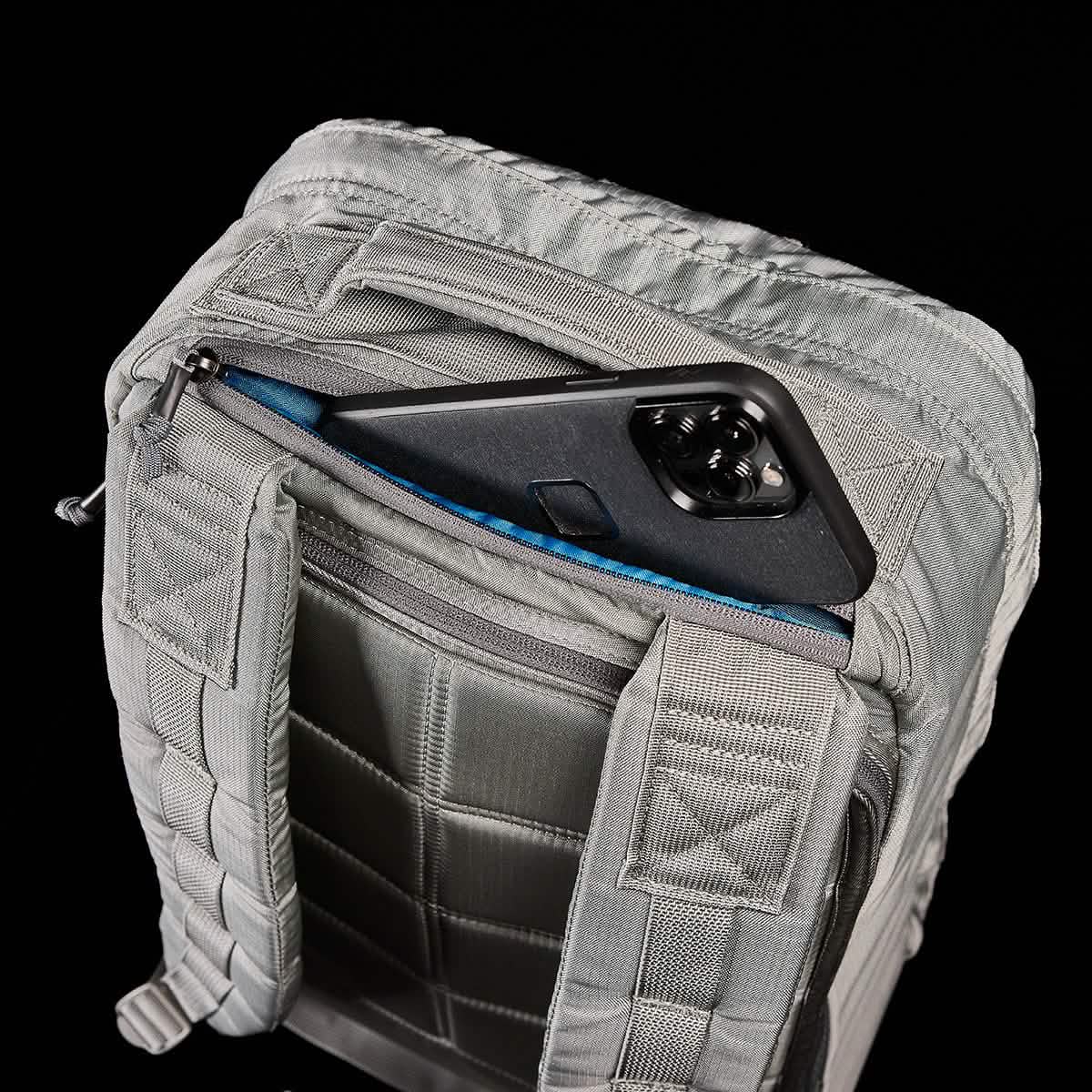 The GR1 USA - Ripstop ROBIC® by GORUCK is displayed with its zipper open, revealing a pocket containing a black smartphone. This ultra-lightweight tactical rucksack features stitched patterns and straps against a black background.