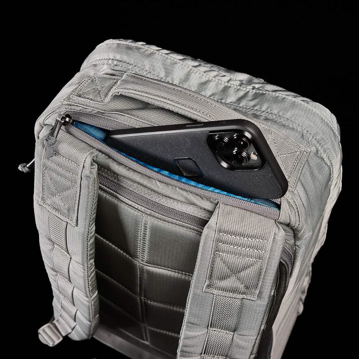 The GR1 USA - Ripstop ROBIC® by GORUCK is displayed with its zipper open, revealing a pocket containing a black smartphone. This ultra-lightweight tactical rucksack features stitched patterns and straps against a black background.