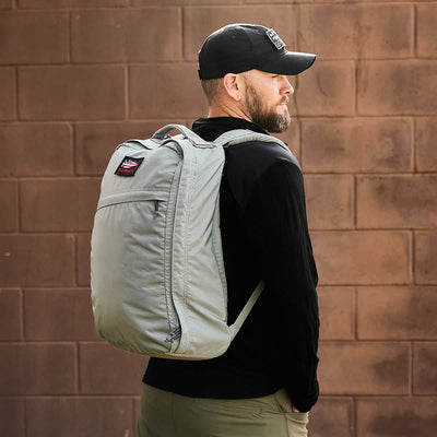 A man stands sideways wearing a black cap and a black jacket, carrying the GR1 USA - Ripstop ROBIC® backpack by GORUCK in ultra lightweight, water-resistant light gray. The background is a brown brick wall.