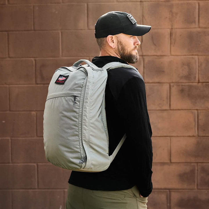 A man stands sideways wearing a black cap and a black jacket, carrying the GR1 USA - Ripstop ROBIC® backpack by GORUCK in ultra lightweight, water-resistant light gray. The background is a brown brick wall.