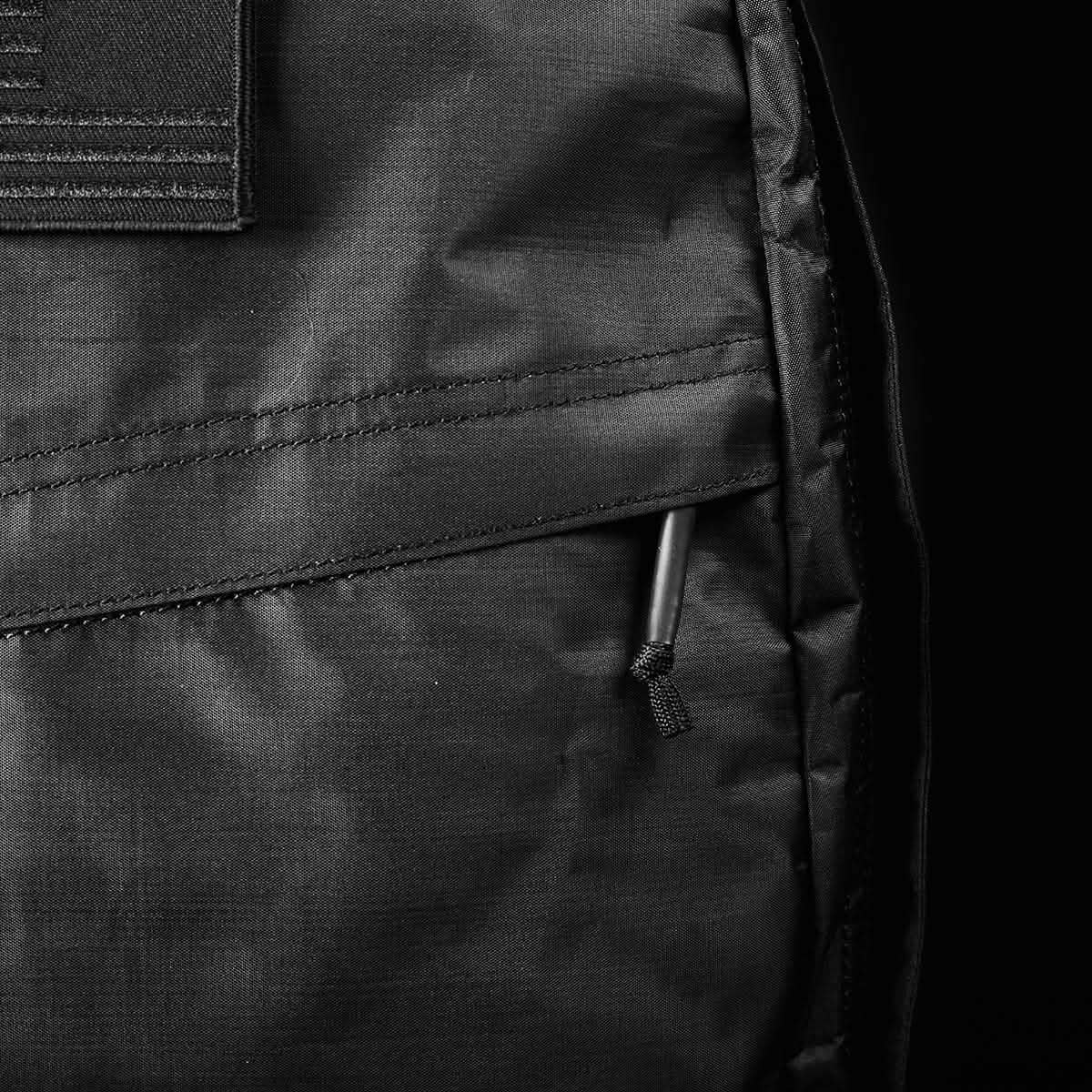 Close-up of a black GORUCK GR1 USA - Dyneema rucksack highlighting a zippered pocket with a corded pull tab. The robust Dyneema® fabric appears smooth, with detailed stitching visible. The monochrome image accentuates texture and craftsmanship.