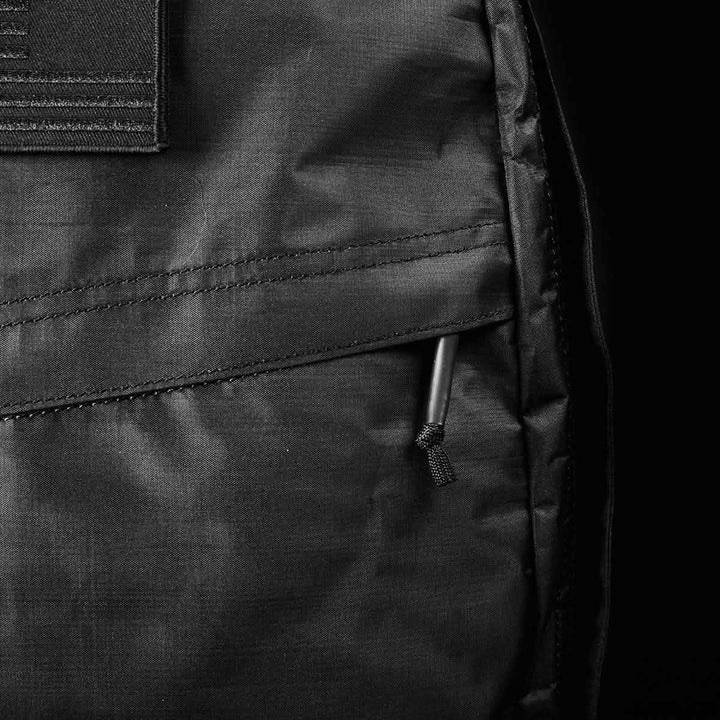 Close-up of a black GORUCK GR1 USA - Dyneema rucksack highlighting a zippered pocket with a corded pull tab. The robust Dyneema® fabric appears smooth, with detailed stitching visible. The monochrome image accentuates texture and craftsmanship.