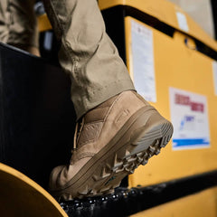 A close-up showcases a person stepping onto a yellow piece of machinery while wearing the MACV-2 Safety Boot - Mid Top by GORUCK. The focus is on the boots and their rugged soles, emphasizing their durability. Their tan pants pair seamlessly with these robust work boots.