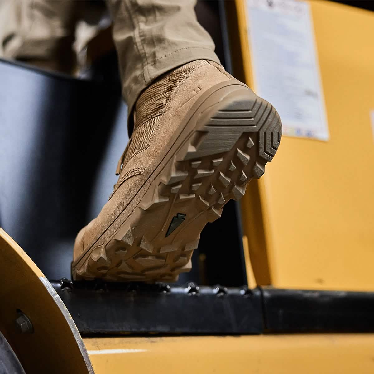 Close-up of a person wearing the GORUCK MACV-2 Safety Boot - Mid Top in tan, featuring rugged soles as they step onto yellow machinery. The focus is on the sole of the safety boot, showcasing its tread pattern. The individual’s lower leg is visible, dressed in beige pants that reflect Special Forces standards in durability and style.