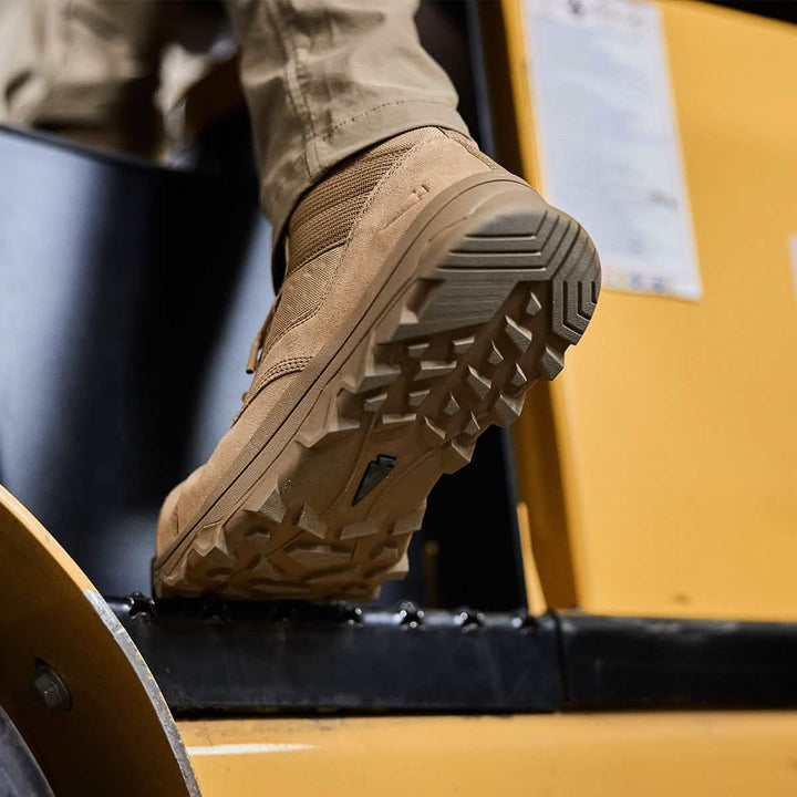 Close-up of a person wearing the GORUCK MACV-2 Safety Boot - Mid Top in tan, featuring rugged soles as they step onto yellow machinery. The focus is on the sole of the safety boot, showcasing its tread pattern. The individual’s lower leg is visible, dressed in beige pants that reflect Special Forces standards in durability and style.
