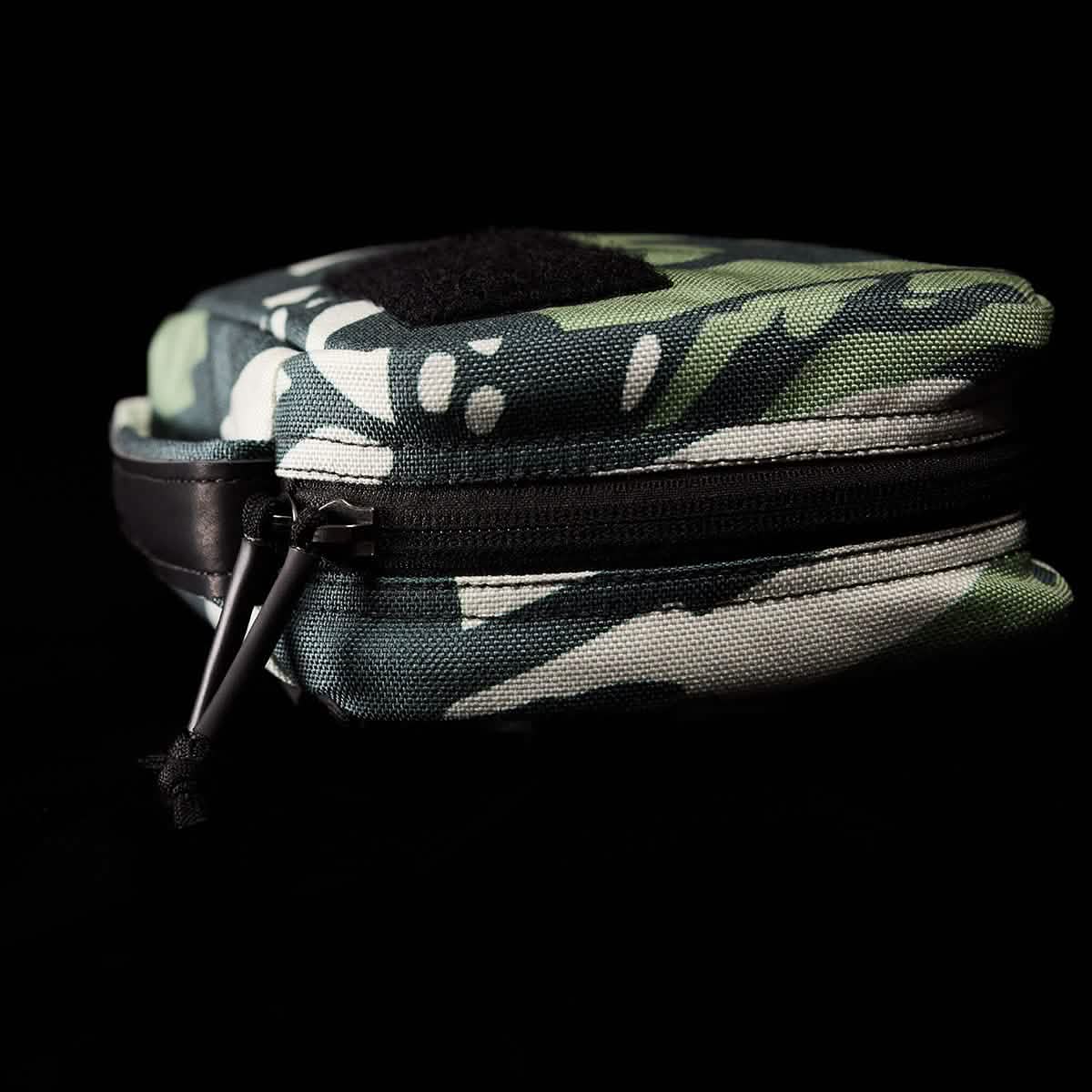 The GR1 Field Pocket x Gear Patrol by GORUCK, a small zippered pouch displaying a Variegated Elephant Ear Camo pattern in green with white spots and black accents, sits against a dark background. The partially unzipped design reveals its sleek black inner lining, perfect for any Gear Patrol adventure.