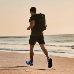 A person, equipped with Men's Rough Runner - White + Speed Grey running shoes from GORUCK, sprints along a sandy beach at sunrise. With a sleek black backpack and dark shorts, they move gracefully as ocean waves gently lap the shoreline in the background.