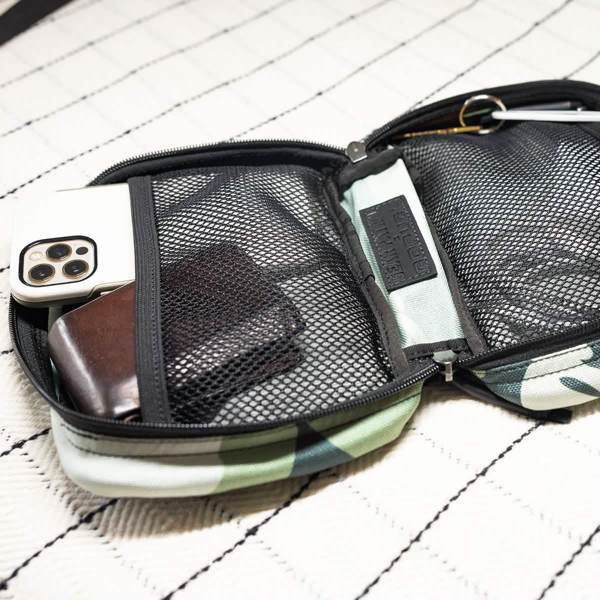 The GR1 Field Pocket x Gear Patrol from GORUCK, in Variegated Elephant Ear Camo pattern, is open on a quilted white surface, revealing a smartphone, a brown wallet, and other small items inside.